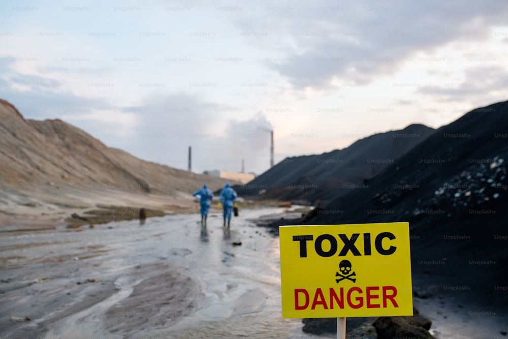 Yellow board with announcement of toxic and dangerous area on background of two contemporary scientists in blue protective coveralls