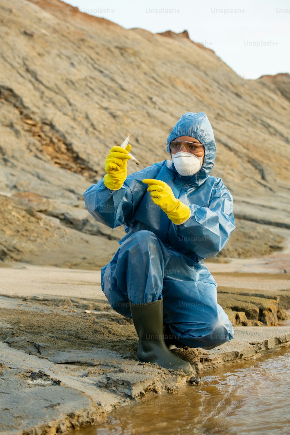 Female ecologist in gloves, coveralls, respirator and eyeglasses looking at flask with sample of dirty water or soil in dangerously polluted area