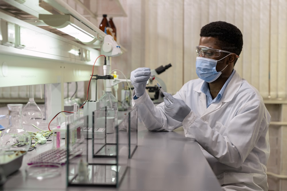 Jovem cientista africano em máscara segurando seringa sentado à mesa no laboratório