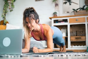 Young woman exercising at home doing push ups and looking at her personal laptop computer to learn or teach workout - content creator business free healthy lifestyle people concept