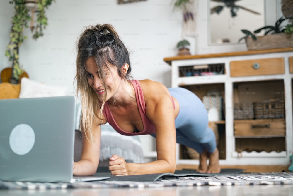 Young woman exercising at home doing push ups and looking at her personal laptop computer to learn or teach workout - content creator business free healthy lifestyle people concept