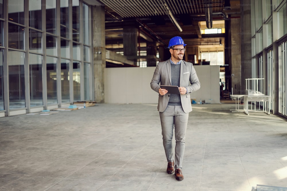 Smiling supervisor walking in building in construction process with tablet in hands and checking on works.