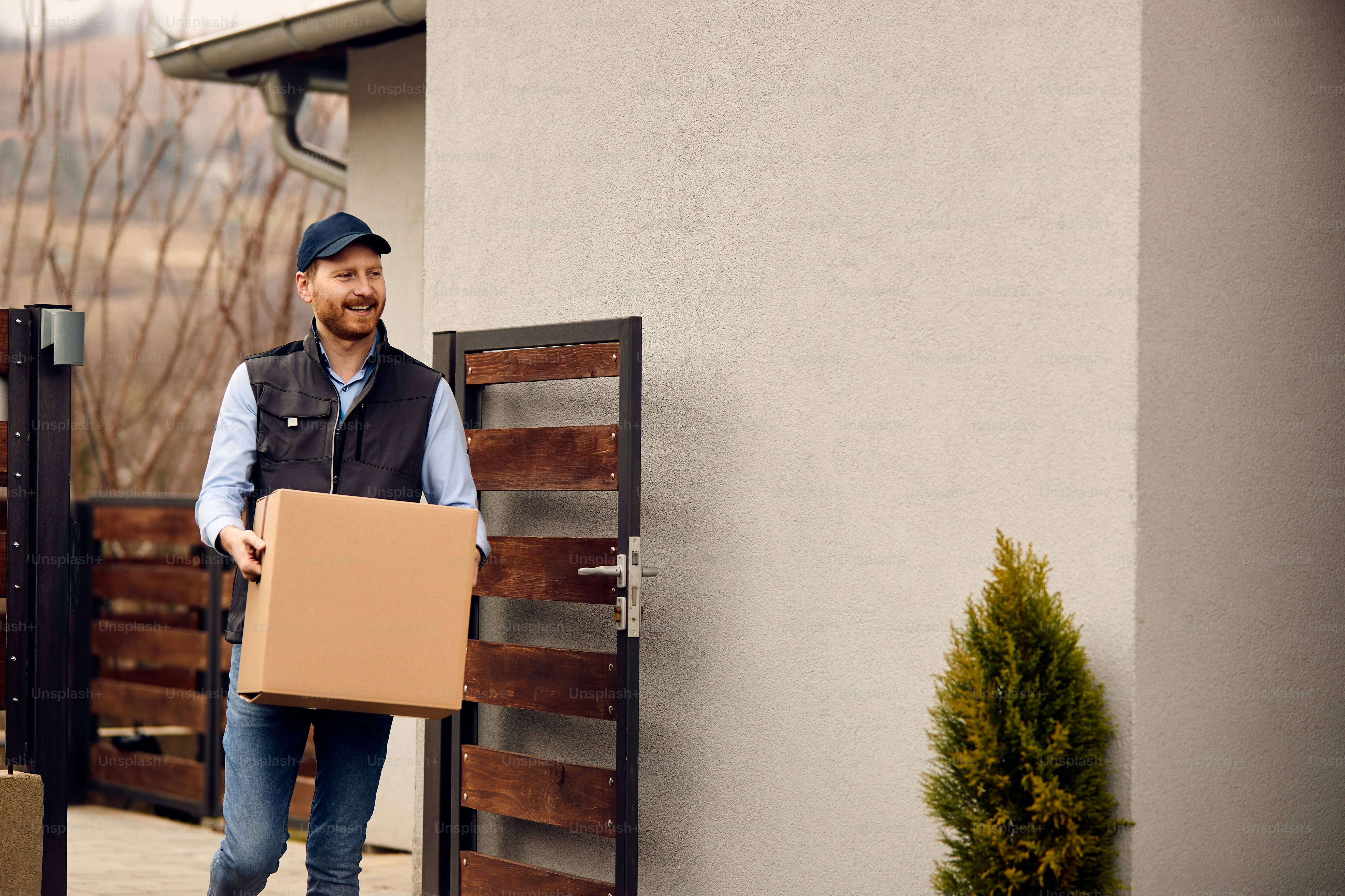 Smiling male courier carrying cardboard box while making home delivery to his customer.