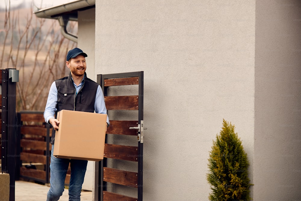 Smiling male courier carrying cardboard box while making home delivery to his customer.