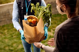 Woman with face mask receiving home delivery and taking paper bag with fresh groceries from delivery person.