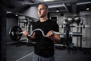 Barbell training. A close-up portrait of half the body of sports and muscular man in the gym exercising his biceps with weights. Modern gym with a dark atmosphere, sports life