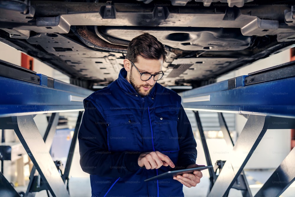 Technical inspection of cars and tablets. A man in uniform holds a tablet in his hands in the workshop canal under the car on a hydraulic elevator. He stands in the canal of the workshop