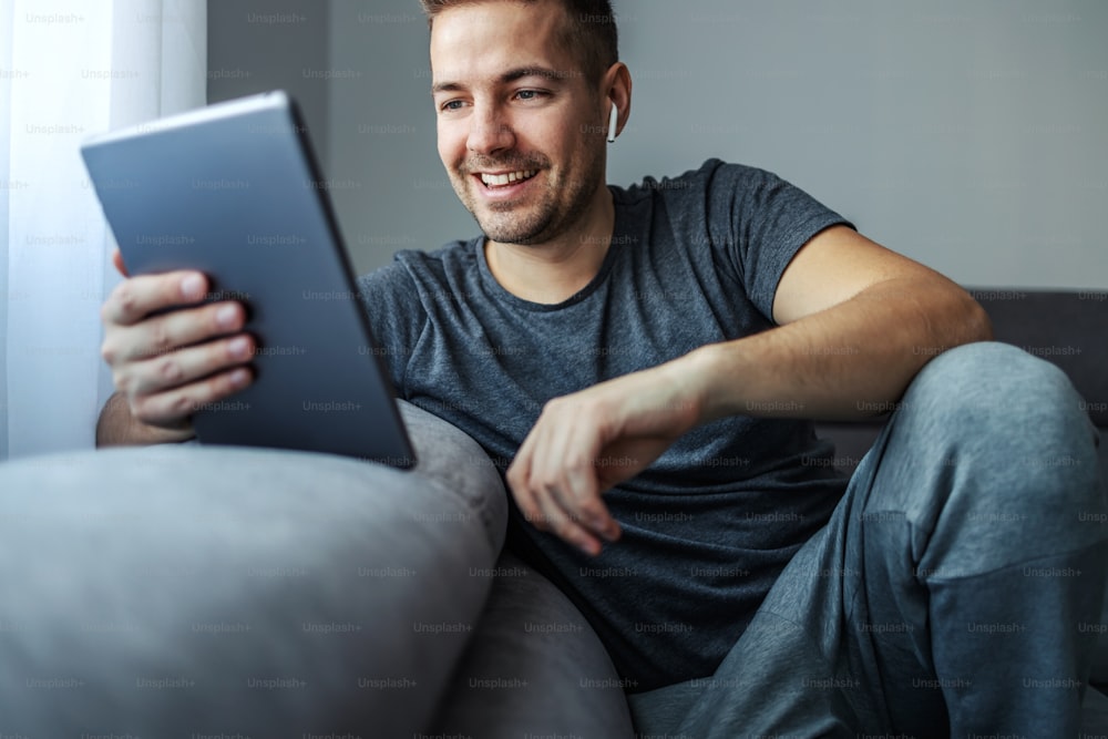 Um homem bonito de meia-idade no conforto e aconchego de casa em roupas esportivas cinza descontraídas senta-se em um sofá na sala de estar do apartamento e olha para algo no tablet que o faz sorrir