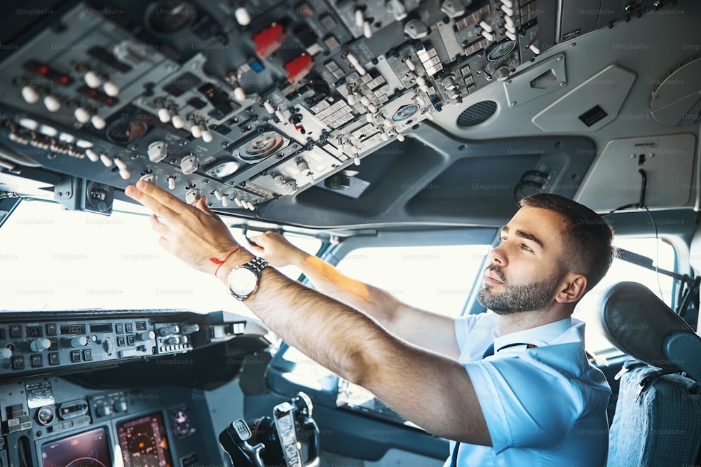 Ruhiger kaukasischer Pilot, der konzentriert aussieht, während er die Umschalter und verschiedene Anzeigen auf dem Armaturenbrett überprüft