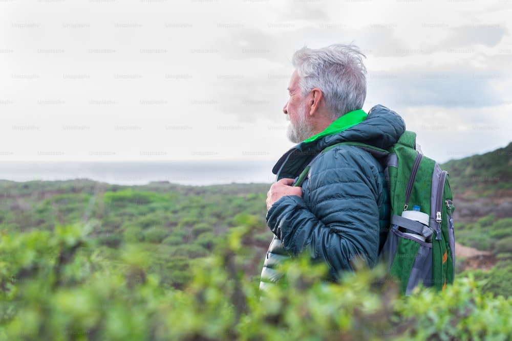Hombre mayor con mochila en excursión al aire libre entre la montaña y el mar en un día lluvioso, mirando el horizonte sobre el agua