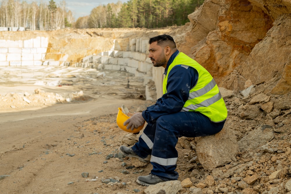 Middle aged builder in uniform sitting on stone while having break