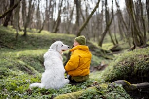 Young woman in hiking clothes sitting near big white dog in green spring forest and looking to each other. Enjoys and explore of tranquil nature.