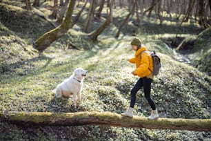 Una mujer joven con ropa de senderismo y mochila pasa tiempo junto con un gran perro blanco en el bosque verde de primavera. Disfruta y explora la naturaleza tranquila. Camina sobre un árbol al otro lado del río.