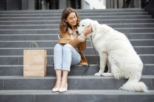 Pretty woman have outdoor lunch near office building with her big white dog while sitting on the stairs. Pet friendly and pet care concept. Animal lover.