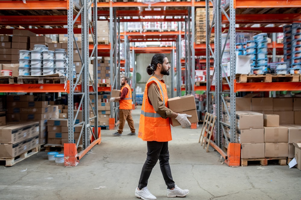 Dark-haired shipping clerk with ponytail and beard carrying boxes over warehouse with goods on shelves