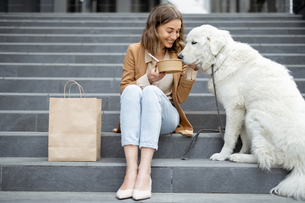 Pretty woman have outdoor lunch near office building with her big white dog while sitting on the stairs. Shares food with a dog. Pet friendly and pet care concept. Animal lover.