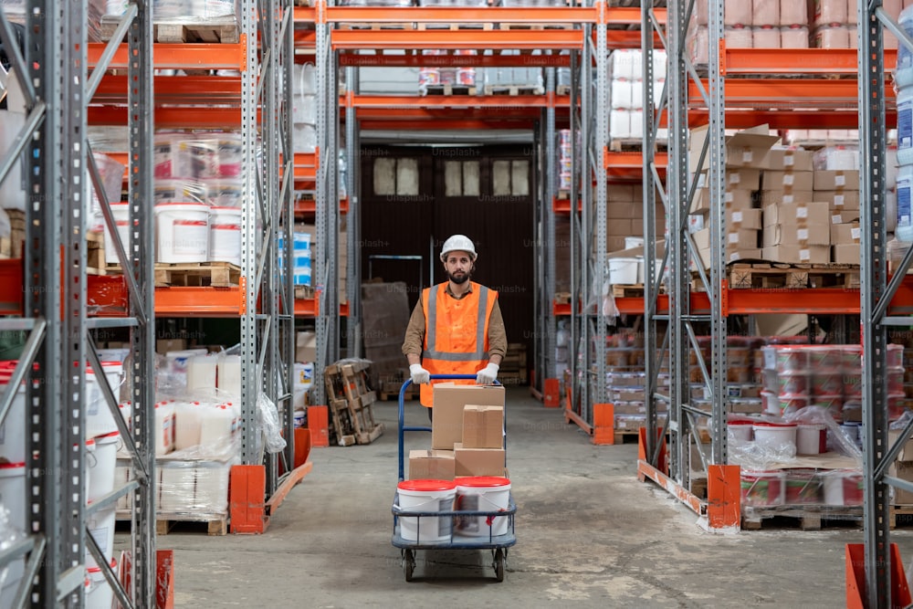 Young mixed race worker in hardhat and orange vest pushing storage cart while sorting boxes at storeroom