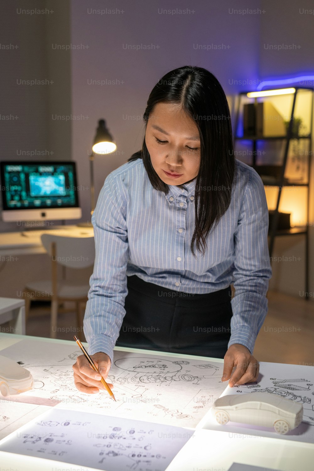 Konzentrierter junger asiatischer Automobildesigner sitzt am Tisch in dunklem Büro und arbeitet mit Layouts von Autokomponenten
