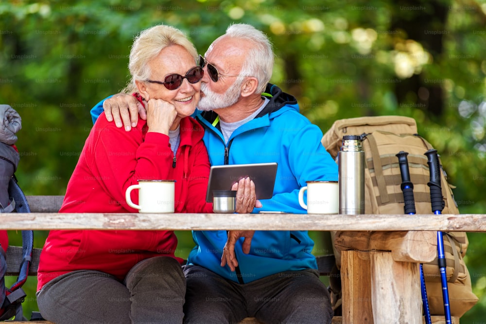 Senior couple hiking. They sit on a bench in the forest ,drink coffee and use tablet.