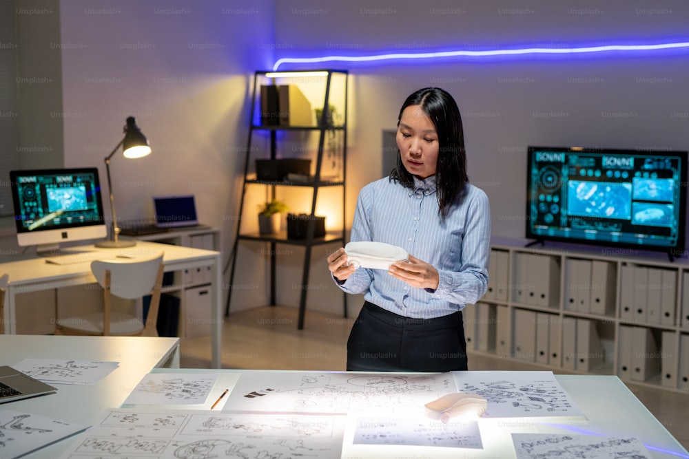 Concentrated young Asian woman in formalwear standing at desk with layouts and holding maquette while producing design of car in office