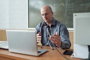 Mature teacher in shirt and eyeglasses sitting by desk, looking at laptop display and talking in microphone during explanation of subject