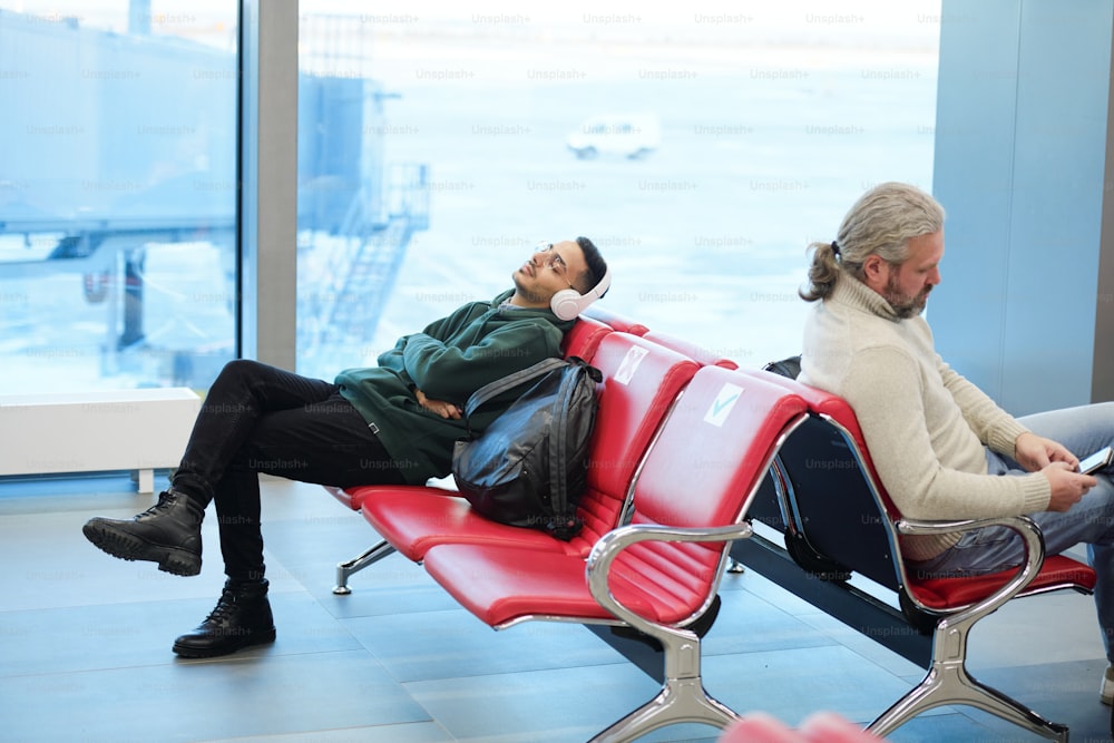 Jovem descontraído com fones de ouvido ouvindo sua música favorita enquanto está sentado no saguão do aeroporto moderno e esperando o voo