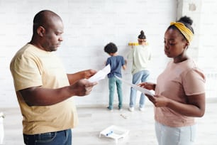 Afrikanischer Mann und Frau in Freizeitkleidung diskutieren Papiere mit Möbelmontageanweisungen, während ihre beiden Kinder an der Wand spielen