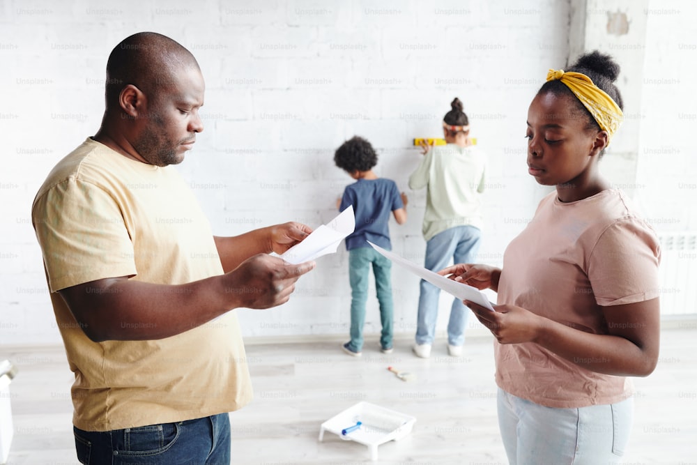 African man and woman in casualwear discussing papers with furniture assembling instructions while their two children playing by wall