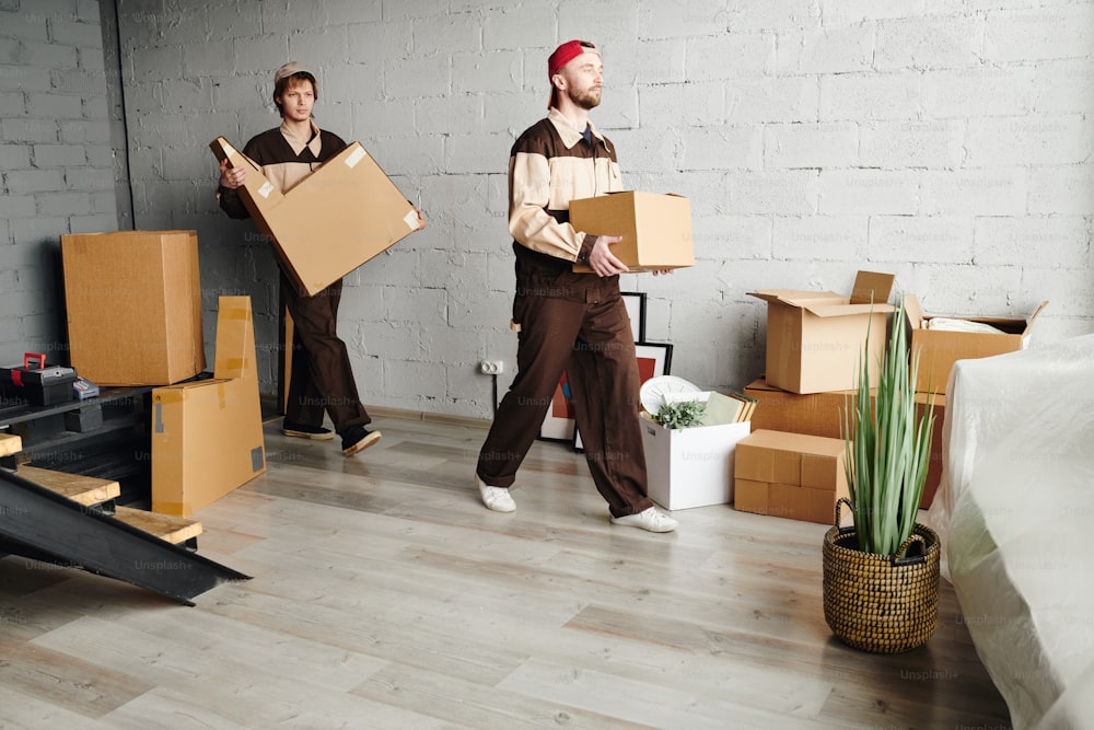 Dois carregadores jovens em trajes de trabalho carregando caixas de papelão embaladas enquanto ajudam a entregar pacotes para novos apartamentos, casas ou estúdios