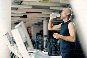 Hombre atlético maduro que toma agua después de trotar en la cinta de correr durante el entrenamiento deportivo en un gimnasio.