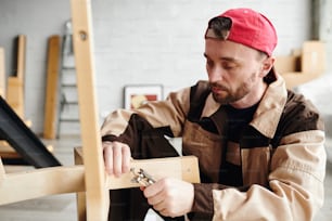 Serious young furniture assemblage specialist in workwear fixing parts of chair while assembling it in large new house, flat or studio