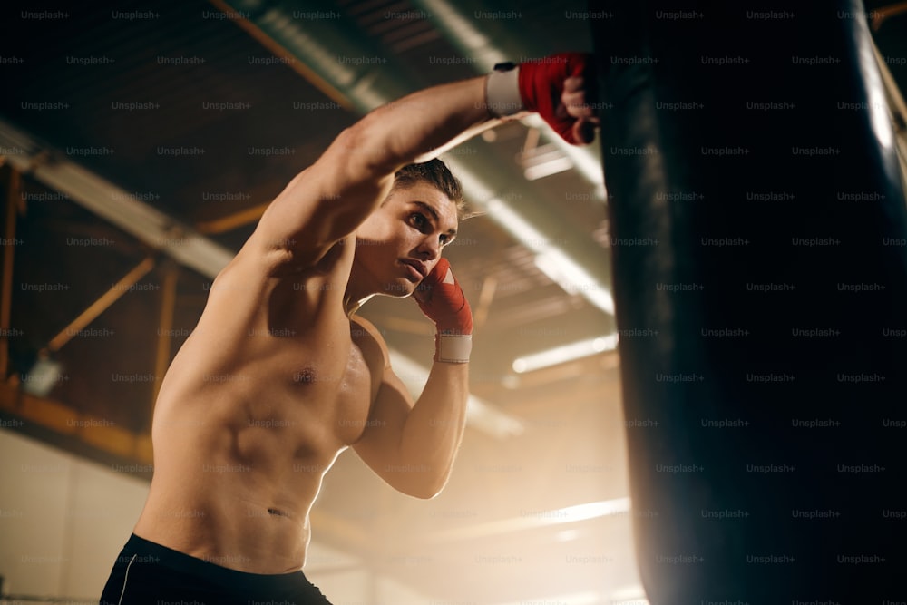 Vista de ángulo bajo de un deportista musculoso que golpea el saco de boxeo mientras hace ejercicio en el club de boxeo.