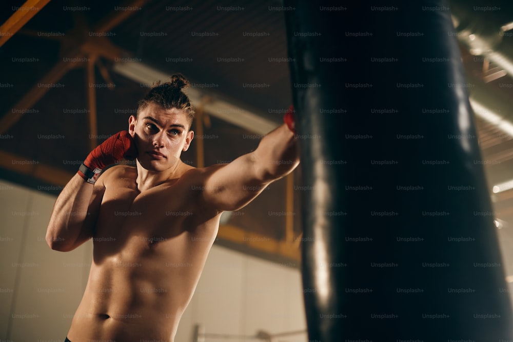 Vista de ángulo bajo de un joven luchador golpeando un saco de boxeo durante el entrenamiento deportivo en un gimnasio.