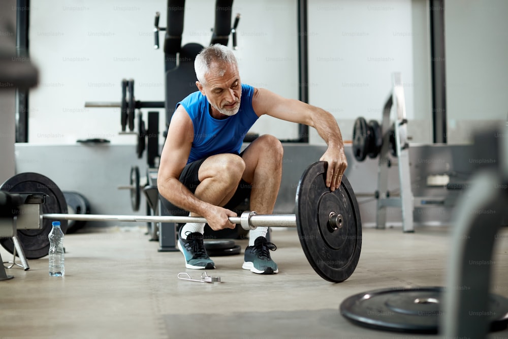 Deportista maduro que ajusta el disco de peso en la barra durante el entrenamiento en el gimnasio.