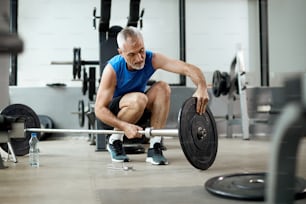 Mature sportsman adjusting weight disk on barbell during gym workout.