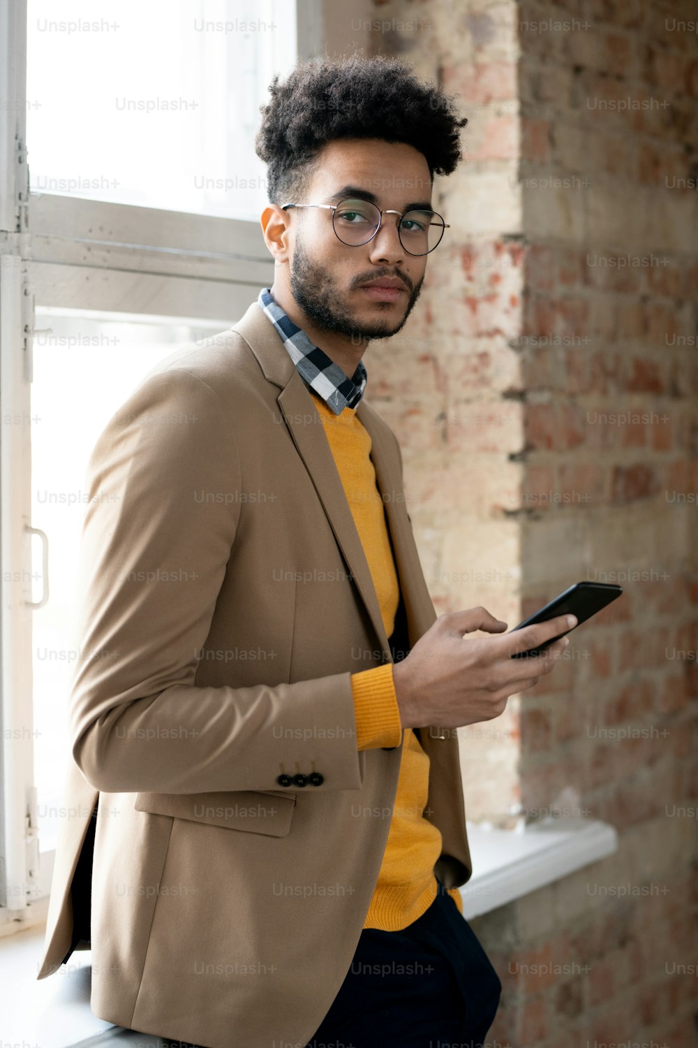 Portrait of serious confident fashionable mixed race businessman standing with gadget at window in corridor of loft style