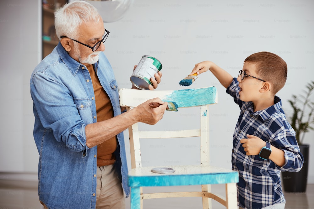 Uomo anziano che si diverte con suo nipote mentre dipinge insieme la sedia a casa.