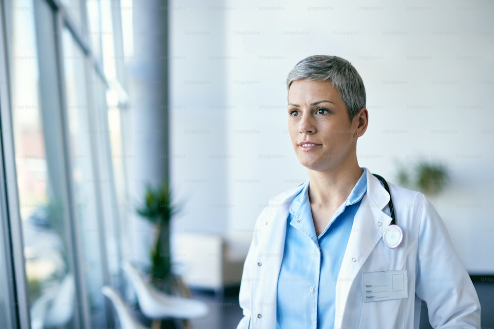 Smiling female general practitioner standing in hospital hallway and looking away.