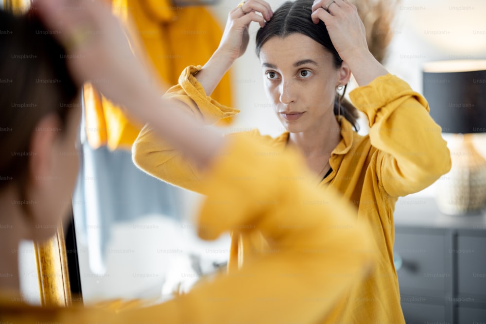 Young woman looks on the mirror and makes hairstyle, getting ready to go out at the hallway at home