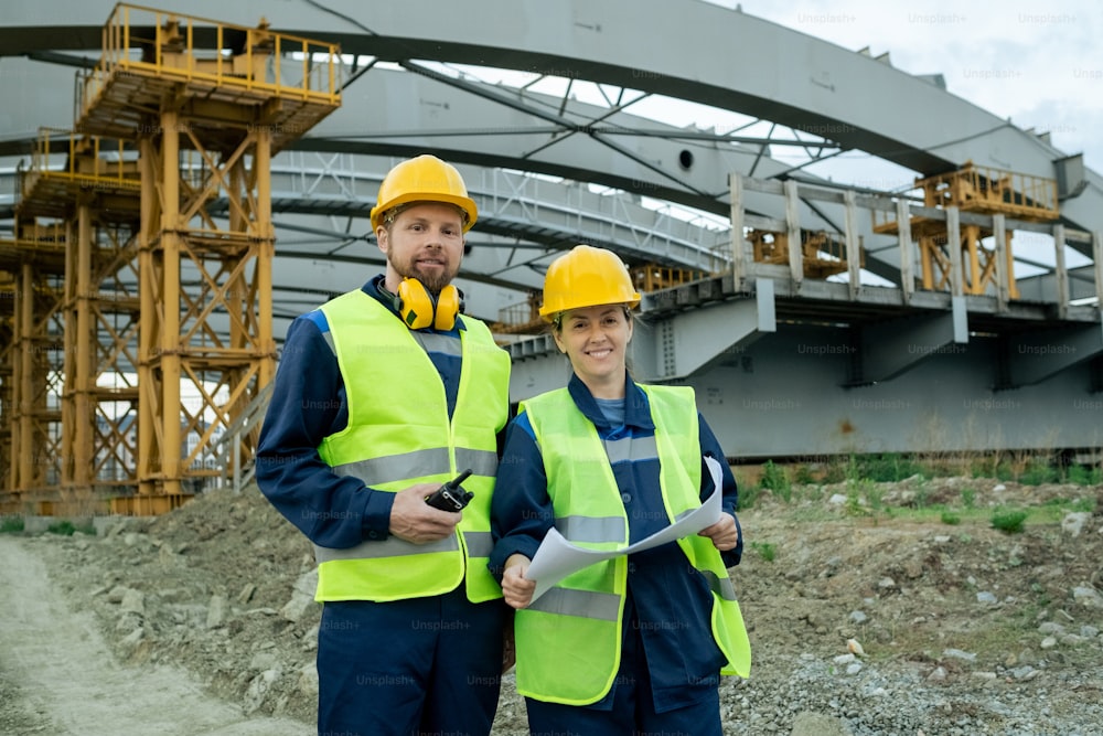 Dos ingenieros exitosos en ropa de trabajo de pie contra un gran marco de construcción