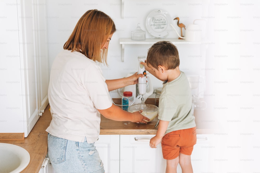 Young woman mother and her toddler boy son cooking with blender at the table in kitchen at home