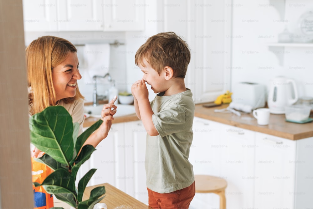 Young woman mother and her toddler boy son have fun while cooking with flour at the table in bright kitchen at home