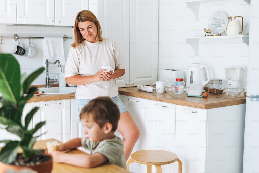 Young woman mother looking as her toddler boy son having breakfast at the table in bright kitchen at home