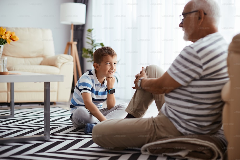Garoto feliz sentado no chão e ouvindo atentamente seu avô que está falando com ele em casa.