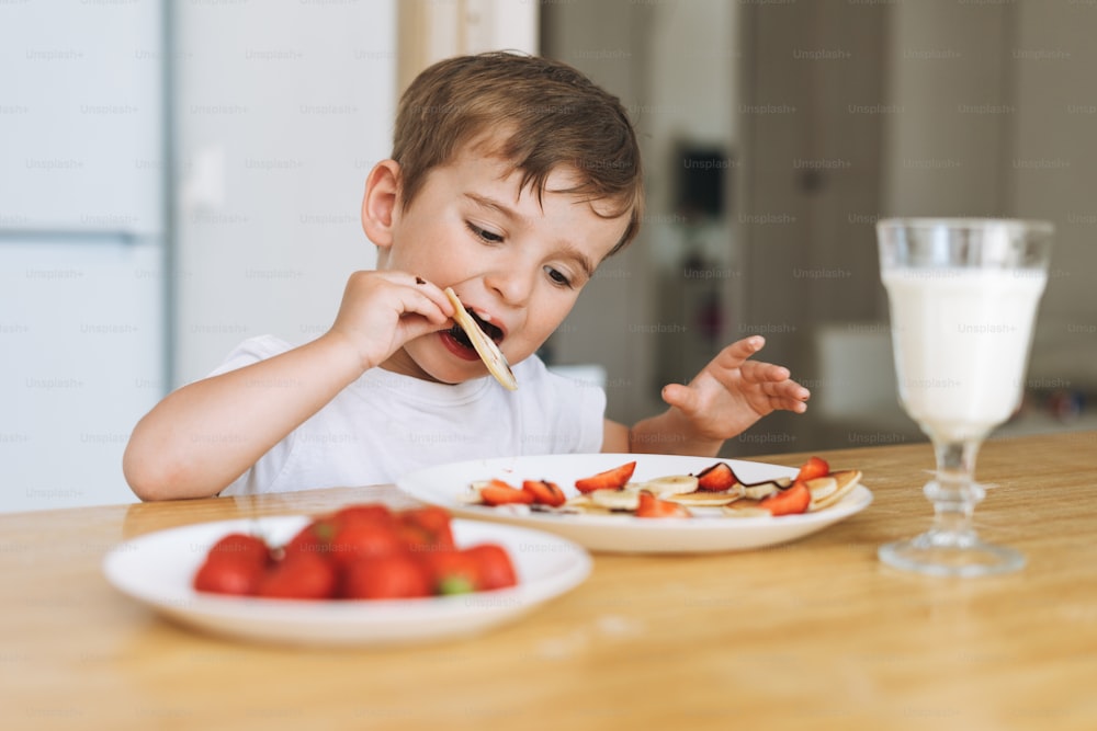 Cute toddler boy having breakfast with puncakes with berries and glass of milk in kitchen at home