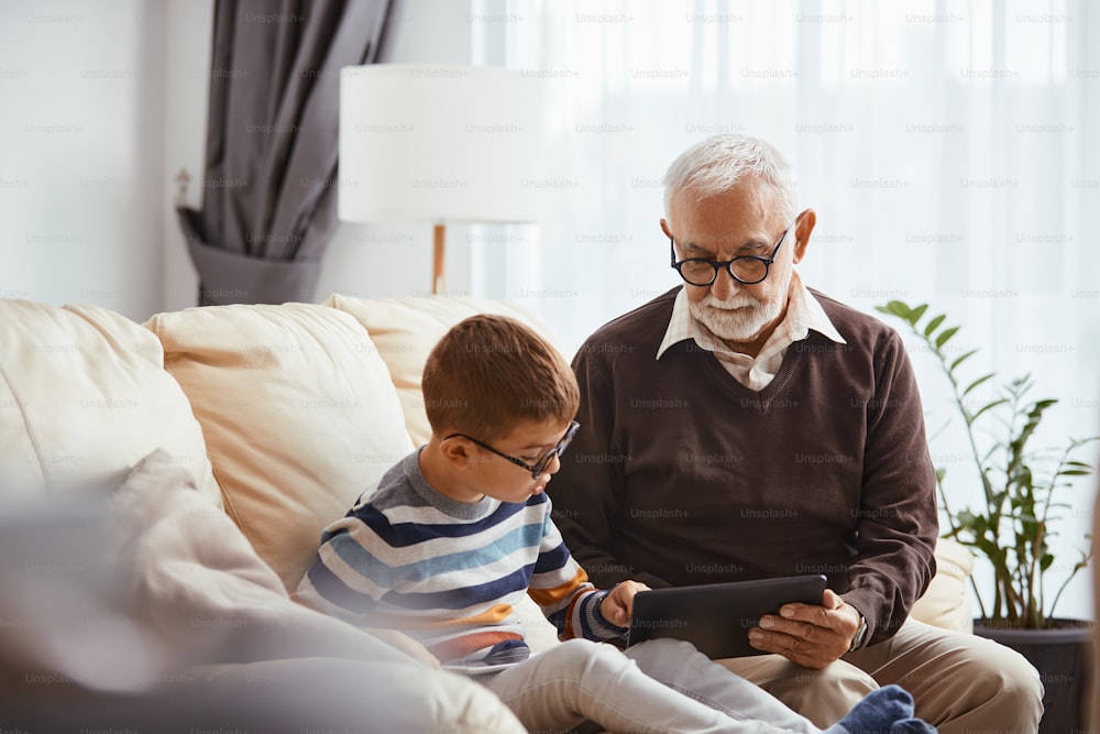 Homem sênior ensinando seu neto a usar touchpad enquanto relaxa no sofá da sala de estar.