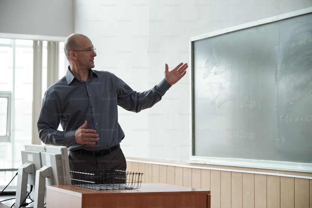 Bald mature teacher looking and pointing at notes on blackboard during explanation