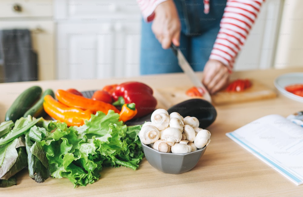 Frisches Gemüse grüner Salat, Paprika, Pilze und Auberginen auf dem Küchentisch. Junge Frau plus Körpergröße positiv in roter Langarmküche mit Buch mit Rezepten auf der Küche