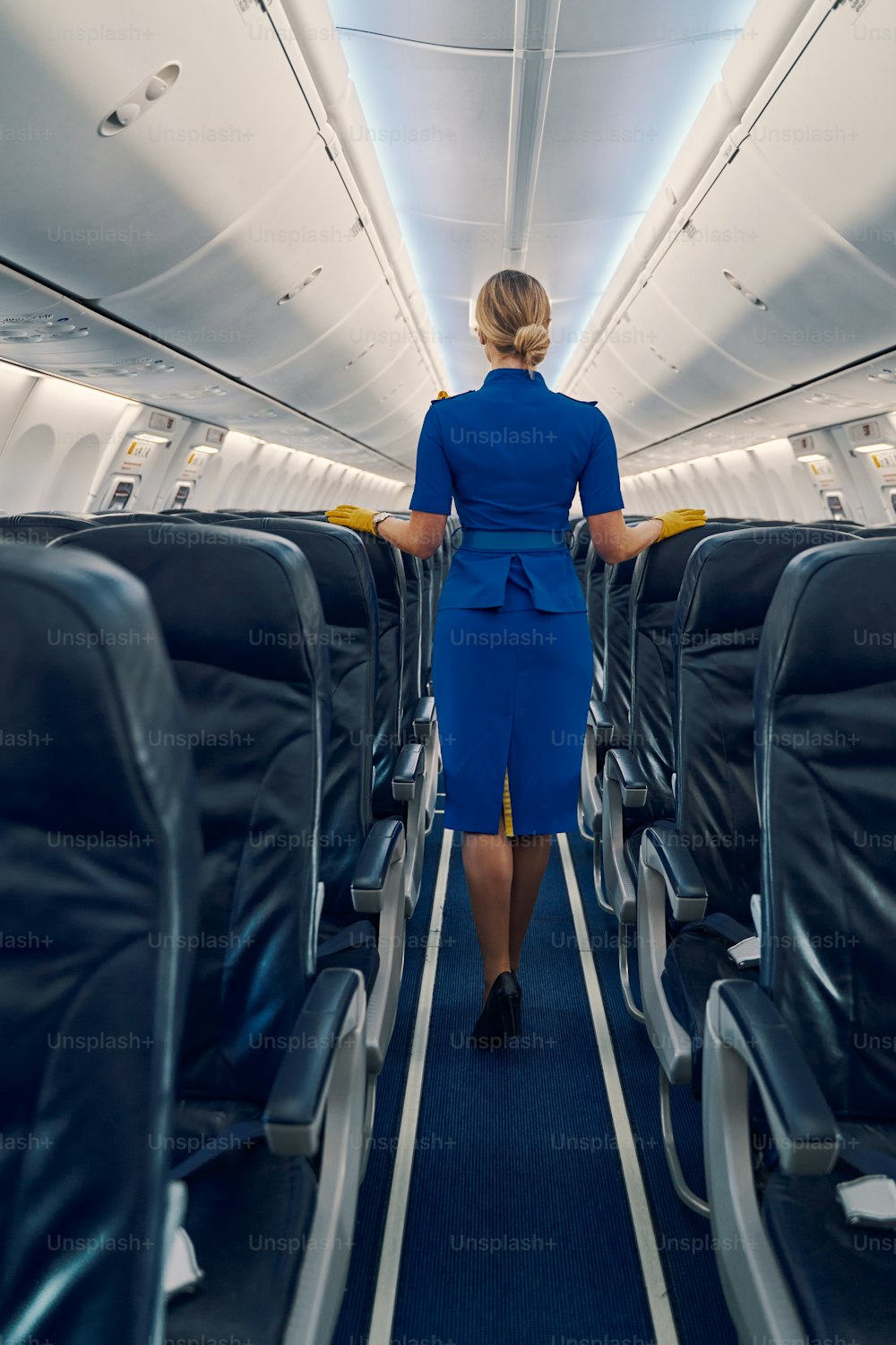 Back view of a slender elegant flight attendant leaning her hands in leather gloves on the airline seats