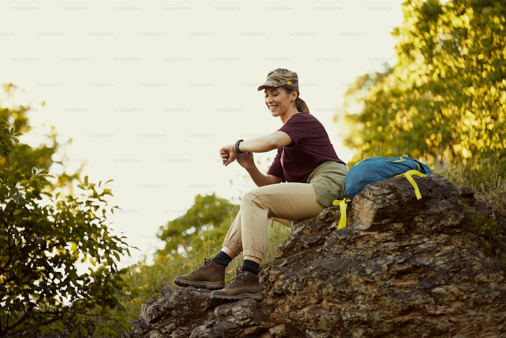 Young happy woman resting on a rock and looking at time on wristwatch while hiking in nature.